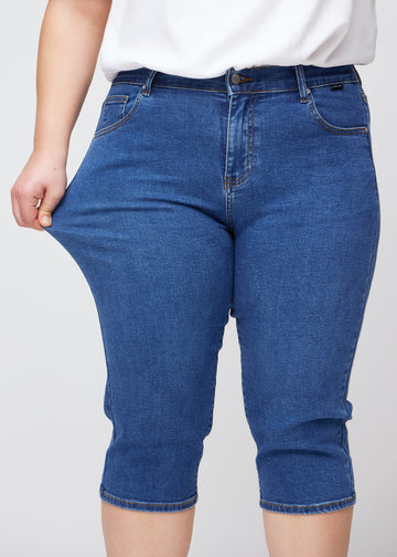 Slim capris for women in dark blue denim, color Oceans. A plus-size model pulls the fabric at the thigh to demonstrate stretch and flexibility.