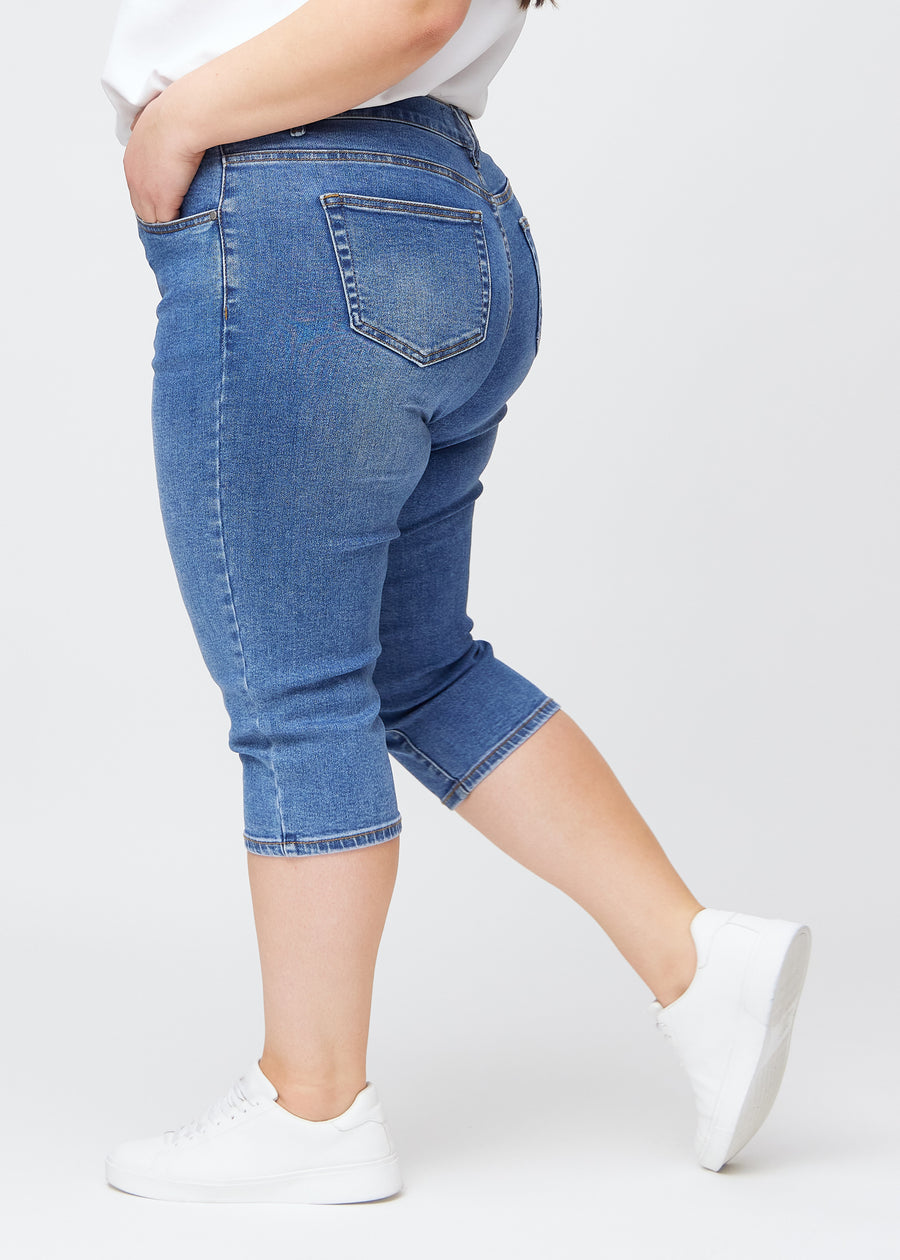 Slim capris for women in blue denim, color Rivers. Side view on a plus-size model, showing the tailored fit and mid-rise waist.