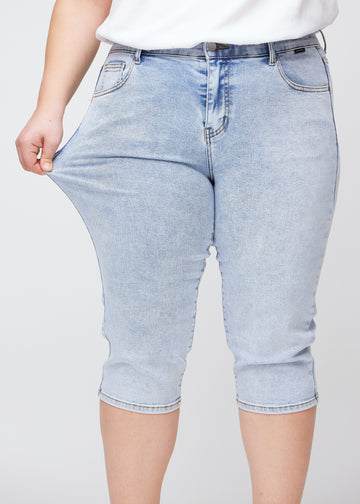 The plus-size model shows the stretch by pulling the capris at the thigh, focusing on comfort and flexibility in the light blue denim.