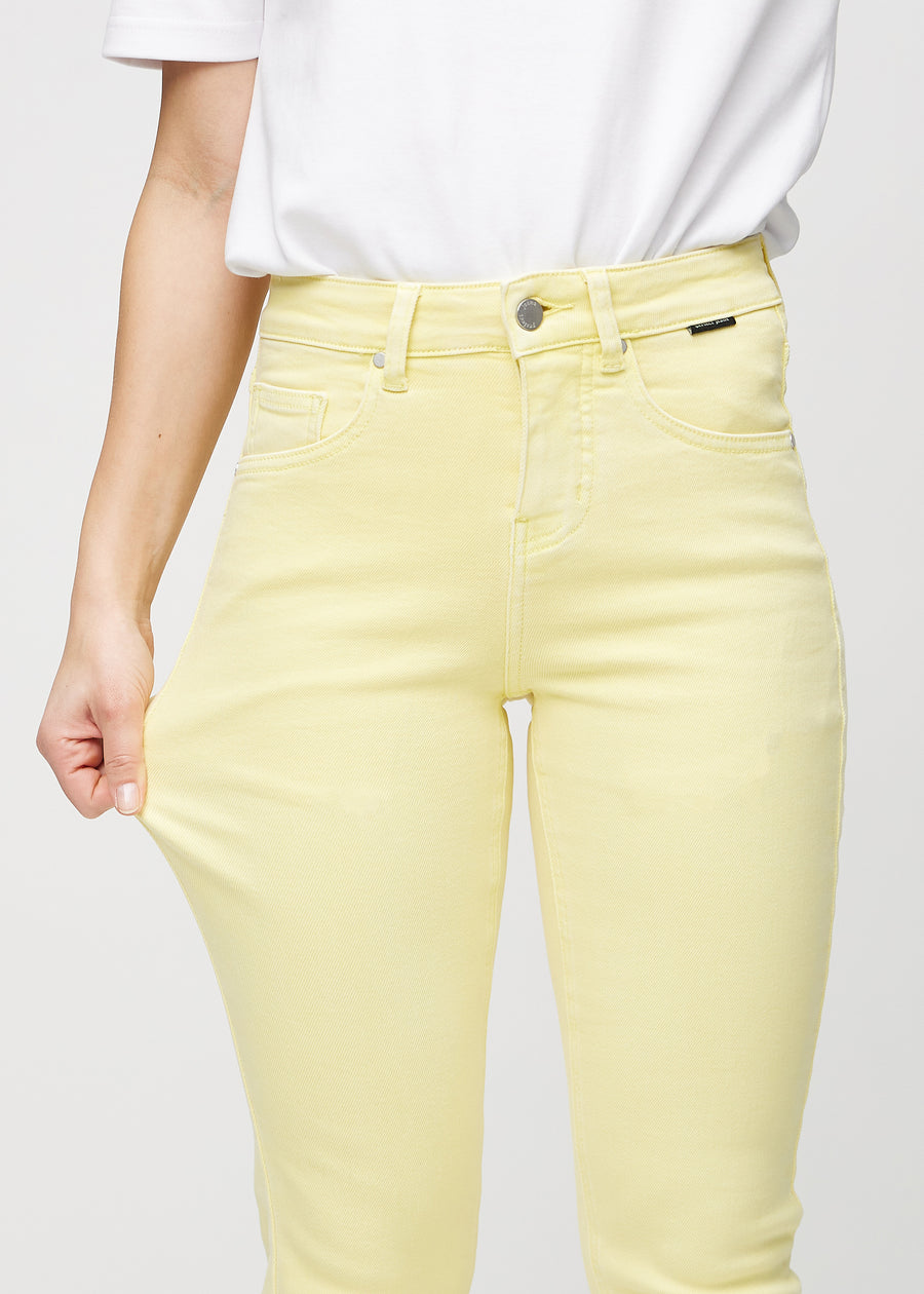 Slim jeans for women in light yellow, color Lemonades. A model stretches the jeans at the thigh to show the fabric&