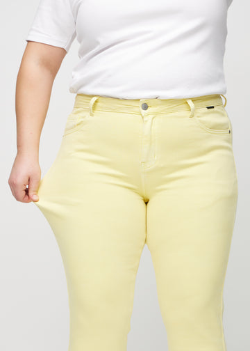 Slim jeans for women in light yellow, color Lemonades. A plus-size model stretches the jeans at the thigh to show the fabric&
