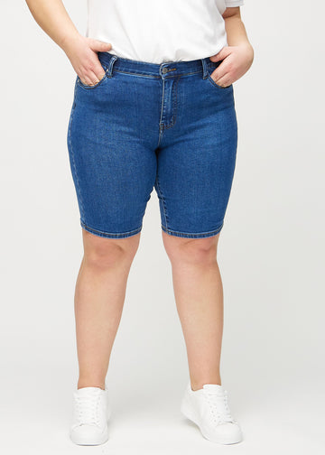 Dark denim skinny middle shorts, model name Oceans, which fit closely around the thigh and end just above the knee on a plus-size model, seen from the front.